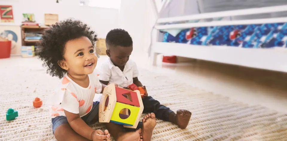 Two toddlers smiling and playing with toys on a classroom rug